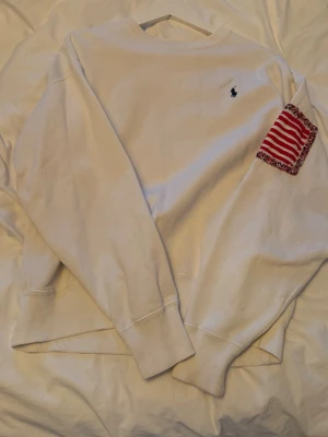 Vit sweatshirt från Polo Ralph Lauren - Vit sweatshirt med blå Polo-tryck på ryggen och liten logga på bröstet. Har en liten fläck brevid märket på fram.