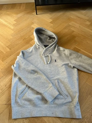 Hoodie från Polo Ralph Lauren - Snygg ljusgrå hoodie från Polo Ralph Lauren med klassisk huva och vita dragsnören. Har en broderad logga i mörkblått på bröstet och en stor magficka framtill. Tillverkad i mjukt bomullsmaterial som känns skönt mot huden. Perfekt för en avslappnad och stilren look.