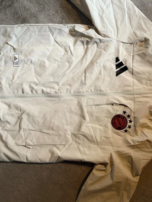 Vit FC Bayern München jacka Adidas - Snygg vit träningsjacka från Adidas med FC Bayern München-logga och svarta detaljer. Jackan har hel dragkedja framtill, sidofickor med dragkedja och klubbmärke broderat på bröstet. Perfekt för fotbollsfans och sportiga tillfällen.
