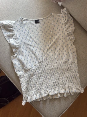 Blommig topp med volangärmar - Säljer denna blus i ljusblå färg med små blommor och volangdetaljer vid ärmarna. Toppen har v-ringning, längst ned vid ringningen har jag sytt för att undvika glapp vid användning! Använd fåtal gånger💕