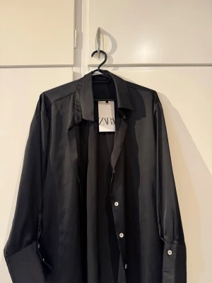 Svart satinliknande skjorta från Zara - Stilren svart skjorta från Zara i satinliknande material. Skjortan har klassisk krage, långa ärmar och vita knappar framtill. Perfekt för dig som gillar en clean och modern look.