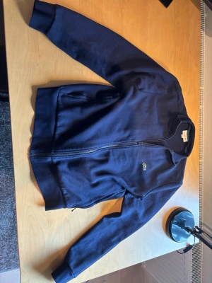 Lacoste hoodie utan luva  - Storlek S/M mer åt M skulle jag säga.Säljer en mörkblå hoodie med dragkedja och långa ärmar. Klassisk design med ribbade muddar och fickor på sidorna. Liten broderad logga på bröstet. Perfekt för dig som gillar enkel och stilren streetwear.