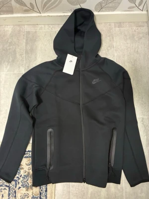 Svart Nike Tech Kofta - Snygg svart hoodie från Nike med hel dragkedja framtill och två fickor med dragkedja. Tröjan har huva och diskret Nike-logga på bröstet. Perfekt för en sportig och avslappnad stil. Materialet känns mjukt och stretchigt.