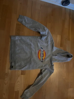 Grå Dickies hoodie med logga - Säljer en grå hoodie från Dickies med stor färgglad logga på bröstet. Hoodien har huva, magficka och ribbade muddar vid ärmslut och nederkant. Perfekt för en avslappnad streetwear-look. Storlek S. Skriv om ni har frågor!!!