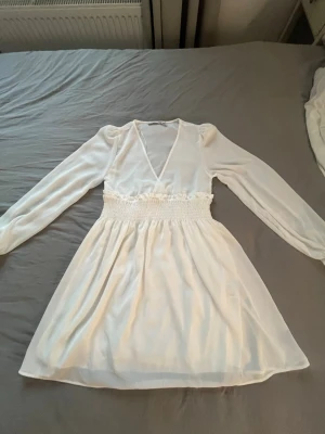 Vit omlottklänning med lång ärm - Söt vit kortklänning med omlott upptill, markerad midja med smock och långa ärmar. Klänningen har en lätt och luftig känsla, perfekt för varmare dagar. Stilren och enkel design som passar till många tillfällen.
