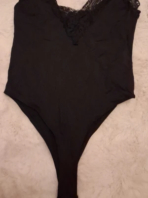 Svart spetsbody med V-ringning - Snygg svart body med djup V-ringning och dekorativ spetsdetalj upptill. Bodyn har en åtsittande passform och är tillverkad i mjukt tyg med stretch. Perfekt att styla under en skjorta eller kavaj för en trendig look.