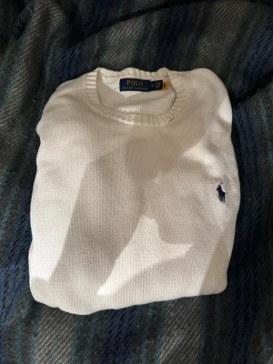 Vit stickad tröja från Polo Ralph Lauren - Klassisk vit stickad tröja från Polo Ralph Lauren med rund halsringning och ribbade muddar. Tröjan har en liten defekt vid halskragen men inget man tänker på. Vid minsta lilla så är det bara att skriva.