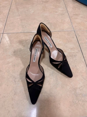 Svarta pumps från Manolo Blahnik - Säljer ett par eleganta svarta pumps från Manolo Blahnik i mocka med spetsig tå och öppen sida. Skorna har smala klackar och dekorativa utskärningar framtill som ger en lyxig och trendig look.