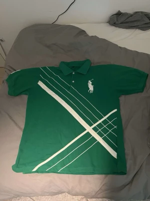 Grön pikétröja med vita ränder - Snygg grön pikétröja med vita diagonala ränder och en stor broderad logga på bröstet. Tröjan har klassisk krage och knappar framtill. Perfekt för dig som gillar sportig stil och vill sticka ut lite extra.