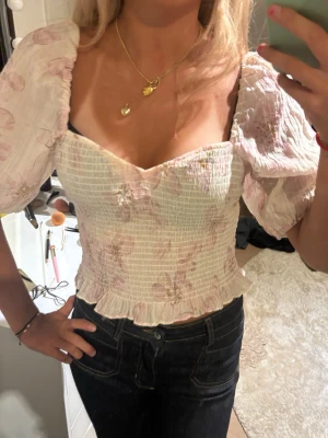 Blommig blus från H&M - Supersöt topp med smockad kropp och volangkant nertill. Toppen är vit med ljusrosa blommigt mönster och har puffiga, korta ärmar. Materialet är tunt och luftigt, perfekt för sommaren. Snygg sweetheart-ringning som ger en romantisk vibe. I storlek M men passar mig som vanligtvis har S