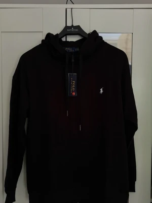 Svart hoodie från Polo Ralph Lauren - Svart hoodie från Polo Ralph Lauren med klassisk huva och dragsko. Broderad vit logga på bröstet och stor känguruficka framtill. Perfekt för en avslappnad och stilren look.