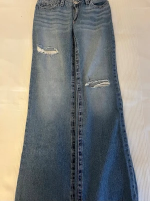 Blå slitna raka jeans - Säljer ett par ljusblå jeans med raka ben och slitningar på båda låren. Jeansen har klassisk femficksmodell, knappgylf och är gjorda i denim. Perfekta för en avslappnad och trendig look. Knappt använda. 