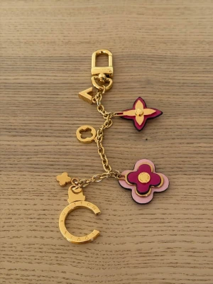  Vuitton guldfärgad väsksmycke - Lyxigt väsksmycke från  Vuitton i guldfärgad metall med detaljer i rosa och lila. Smycket har flera ikoniska LV-blommor och en halvmåneformad berlock. Perfekt att hänga på väskan eller använda som nyckelring för en exklusiv touch.
