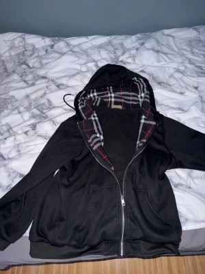 Svart hoodie med rutigt foder - Säljer en svart hoodie med dragkedja från Burberry. Insidan av huvan har det klassiska Burberry-rutiga fodret i svart, vitt och rött. Hoodien har fickor framtill och justerbar huva med snören. Perfekt för dig som gillar stilrena detaljer och ikoniskt mönster.