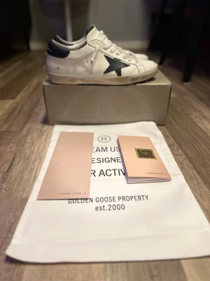 Golden Goose  - Golden Goose sneakers använda kanske 9 gånger säljer för att dom inte passade mig så bra. Det är i vitt läder med marinblå stjärndetalj på sidan och marinblå häl. Klassisk låg modell med vita skosnören och slitna detaljer för en trendig look. Kommer med originalpåse och tillbehör. innåt är 28cm längd och 9cm bredd inutti