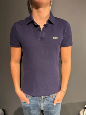 Marinblå pikétröja från Lacoste - Klassisk marinblå pikétröja från Lacoste med korta ärmar och krage. Tröjan har den ikoniska krokodilloggan på bröstet och knappar vid halsen. Perfekt till jeans eller shorts för en clean och stilren look. Modellen är 175cm och väger 70kg