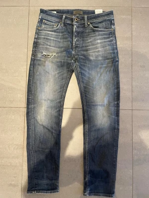 Blå slitna jeans med smal passform - Säljer ett par blå jeans från Jack & Jones med smal passform och snygga slitningar på låret. Jeansen har klassisk femficksdesign, dragkedjegylf och ljusa tvättade partier för en trendig look. Perfekta för dig som gillar en avslappnad men stilren stil.