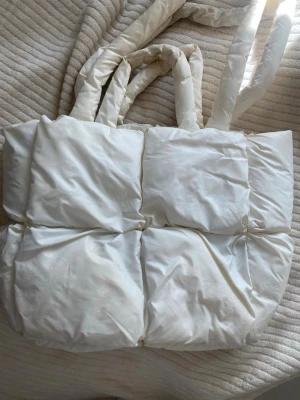 Vit quiltad puffig handväska - Trendig vit handväska med quiltad, puffig design och breda axelband. Väskan har en mjuk, vadderad look och är tillverkad i tygmaterial. Perfekt för dig som gillar en unik och cozy stil. Rymlig insida och enkel att matcha med olika outfits.