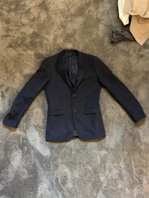 Mörkblå kostym med kavaj och byxor/ Bläck  - Snygg mörkblå kostym från bläck med klassisk tvåknäppt kavaj och matchande byxor. Kavajen har smal passform, två fickor med lock och en bröstficka. Byxorna är raka och har bälteshällor. Perfekt för dig som vill ha en stilren och tidlös look.