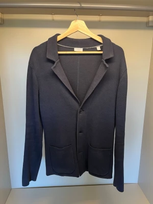 Stickad Kavaj - Stilren mörkblå stickad kavaj med klassisk krage och två stora framfickor. Modellen har en knappstängning framtill och är tillverkad i ett mjukt, stickat material. Perfekt för dig som gillar en enkel men snygg look.