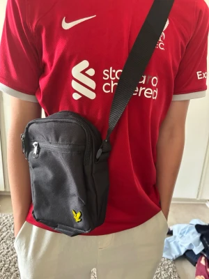 Svart axelväska med gul logga - Snygg svart axelväska från Lyle & Scott med justerbar rem och gul broderad logga framtill. Väskan har ett huvudfack med dragkedja och en mindre ytterficka, perfekt för mobilen och andra småprylar. Tillverkad i slitstark textil.