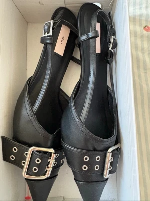 Svarta pumps  - Snygga svarta pumps med spetsig tå, breda remmar och stora silvriga spännen. Skorna har tunna ankelremmar och är tillverkade i konstläder. Klackhöjd 4,5 cm. Storlek 38 i bra skick🖤