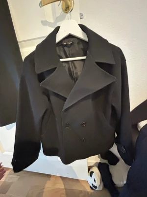 Svart kort kappa med bred krage från Chelsea - Stilren svart kappa med dubbelknäppning och bred krage. Kortare modell som ger en modern look. Perfekt för kyliga dagar och enkel att matcha med olika outfits. Kappan har slät yta och är fodrad för extra komfort.