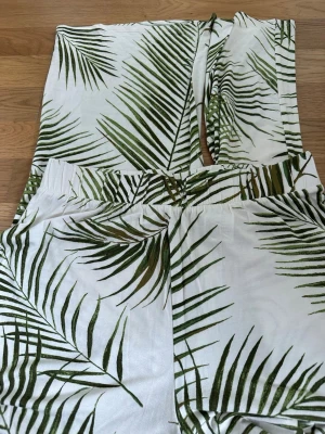 Vita linnebyxor med palmbladsmönster - Säljer ett par vita linnebyxor med grönt palmbladsmönster. Byxorna har rak passform och resår i midjan för extra komfort. Perfekta för en avslappnad och trendig look under varma dagar. Materialet känns svalt och luftigt.