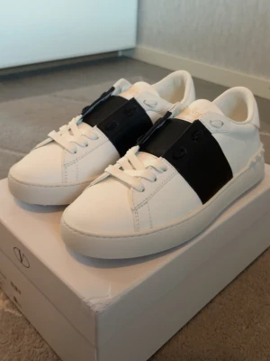 Valentino Garavani vita sneakers - Snygga vita sneakers från Valentino Garavani med bred svart läderdetalj över snörningen. Klassisk låg modell med rund tå och vita sulor. Tillverkade i slätt läder med diskret logga på plösen. Perfekta för en stilren och trendig look.