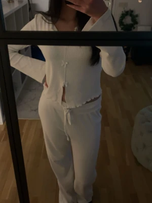 Vit ribbad pyjamas med rosetter - Mysig vit pyjamas i ribbat material med långärmad topp och byxa. Toppen har små rosetter framtill och vågig kant nedtill. Byxorna har dragsko i midjan för skön passform. Perfekt för chillkvällar hemma.