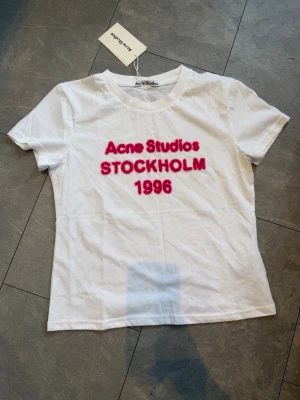 Vit Acne Studios t-shirt med tryck - Vit t-shirt från Acne Studios med rosa texttryck 'Acne Studios STOCKHOLM 1996' på bröstet. Klassisk rund halsringning och korta ärmar. Perfekt basic med en twist för dig som gillar stilrena plagg med statement.