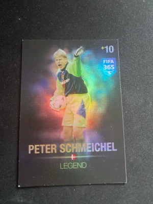 FIFA 365 Peter Schmeichel Legend-kort - Samlarobjekt: ett holografiskt FIFA 365 Panini Adrenalyn XL-kort med Peter Schmeichel. Kortet har färgglad bakgrund, texten 'LEGEND' och visar Schmeichel i målvaktsställning med boll. Perfekt för fotbollsfans och samlare.