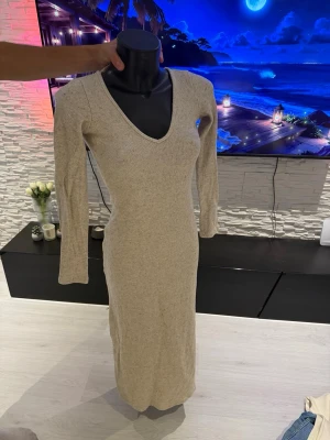 Beige stickad långklänning - Mysig beige stickad långklänning med v-ringning och långa ärmar. Klänningen har en figurnära passform och är perfekt för dig som gillar en enkel men stilren look. Passar till både sneakers och boots.