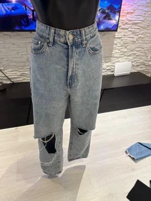 Ljusblå mom jeans med stora hål - Säljer ett par ljusblå mom jeans med hög midja och stora slitningar på båda benen. Jeansen har klassisk femficksdesign och raka ben. Perfekta för en avslappnad och trendig look. Materialet är denim i bomull.