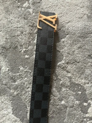 Svart rutigt bälte  - Snyggt bälte i svart och grått med rutigt mönster och metallspänne. Bältet har flera hål för justering och en stilren design som passar till många olika outfits. 116cm