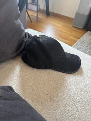 Svart keps med diskret mönster - Snygg svart keps med diskret ton-i-ton mönster och böjd skärm. Kepsen har en liten metallknapp på toppen och justerbar rem bak. Passar perfekt till en streetwear-look eller för att lyfta en enkel outfit.