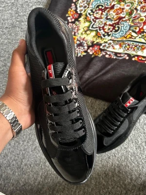 Svarta p cups  - Snygga svarta sneakers med glansiga lackdetaljer och meshpaneler. Skorna har svarta skosnören, metallhål och en röd detalj på plösen. Tidlös design som passar till det mesta och ger en clean look.