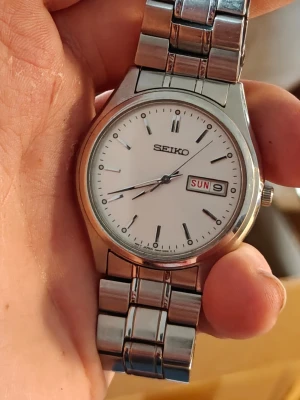 Seiko silverfärgad klocka - Klassisk Seiko armbandsklocka i silverfärgat stål med vit urtavla och dag- och datumvisning.Inställning av Dag fungerar ej. Klockan har stilrena indexmarkeringar och ett robust metallarmband som ger en tidlös look.