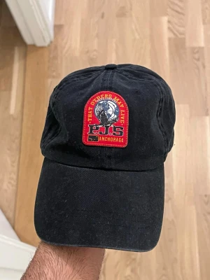 Svart keps med patch och text - Säljer en svart keps med böjd skärm och justerbar rem bak. Framtill sitter en färgglad patch med texten 'THAT OTHERS MAY LIVE' och 'PJS ANCHORAGE' i rött och gult, samt en bild på en örn. Kepsen är i bomull och har en avslappnad passform.