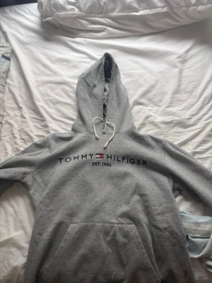 Grå hoodie från Tommy Hilfiger - Säljer en grå hoodie från Tommy Hilfiger med klassisk logga och text 'EST. 1985' på bröstet. Hoodien har huva med vita snören och en stor magficka. Materialet är mjukt och bekvämt, perfekt för chill dagar.