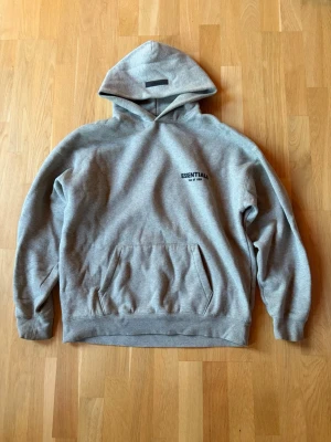 Grå Essentials Fear of God hoodie - Säljer en grå hoodie från Essentials Fear of God med svart logga på bröstet och stor text på ryggen. Klassisk känguruficka framtill och huva. Tillverkad i mjukt sweatshirtmaterial.