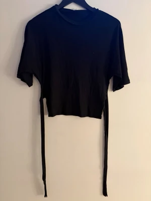 Svart croppad topp med knytband - Snygg svart croppad topp med korta ärmar och långa knytband i sidorna som kan knytas i ryggen för en cool detalj. Perfekt till höga jeans eller kjol. Enkel och stilren design som passar till mycket.