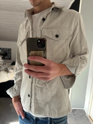 Overshirt  - Snygg beige overshirt i manchestertyg med två bröstfickor och svarta knappar. Rak passform och klassisk krage, perfekt att bära över en t-shirt. Ärmarna kan vikas upp för en mer avslappnad look.