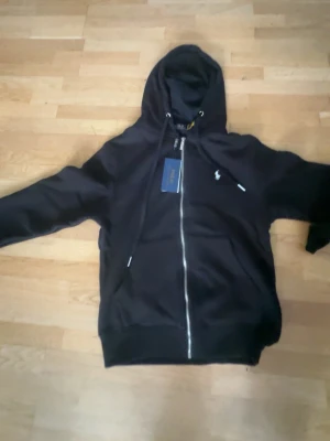 Ralph lauren zip-hoodie - Säljer en svart hoodie med dragkedja och huva. Hoodien har dragsko i huvan, fickor framtill och en liten vit logga på bröstet. Tillverkad i mjukt material som passar perfekt till en avslappnad stil.