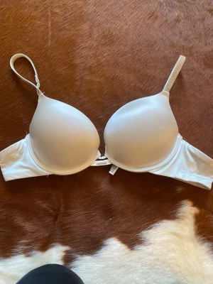 Vit push-up bh med strassdetalj - Snygg vit push-up bh med släta kupor och justerbara axelband. Bh:n har en liten strassdetalj mellan kuporna som ger en extra touch. Materialet är glansigt och känns mjukt mot huden. Perfekt för dig som vill ha en enkel men elegant bh. Aldrig använd 