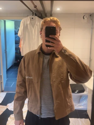Brun workwear jacka med dragkedja - Säljer en brun workwear-inspirerad jacka i kraftigt bomullstyg. Jackan har dragkedja framtill, två stora bröstfickor och en klassisk krage. Detaljerad med broderad text på bröstet och patch bak. Även croppad 