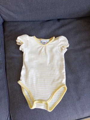 Gulrandig body från Kaxs by KappAhl - Supersöt body i mjuk bomull från Kaxs by KappAhl. Den har ljusgula och vita tunna ränder, puffärmar och ett litet broderat hjärta framtill. Kantad med gul ribb runt hals, ärmar och ben, samt tryckknappar i grenen för enkel påklädning.