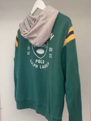 Ralph Lauren hoodie - Vintage Ralph Lauren hoodie med grå huva från Polo Ralph Lauren. Stora RL-bokstäver på bröstet och vita tryck på ryggen. Passar 160-170cm. 