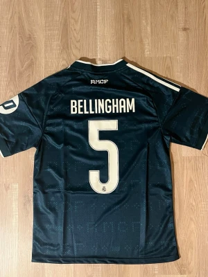 Real Madrid Bellingham bortatröja 26/27 Mörkgrön - Snygg mörkgrön Real Madrid bortatröja med Bellingham och nummer 5 på ryggen. Tröjan har vita detaljer, klubbmärke på bröstet samt Emirates Fly Better som sponsortryck. Tillverkad i ett lätt och ventilerande material, perfekt för fotboll. HELT NY!