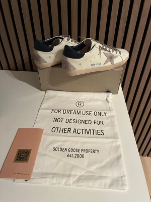 Golden Goose vita sneakers stjärna - Golden Goose sneakers i vitt läder med beige mockadetaljer, grå snörning och en ikonisk stjärna i metallic på sidan. Mörkblå hälkappa och texten 'GOLDENSTAR' på sidan. Snygg vintage-look och klassisk låg modell.